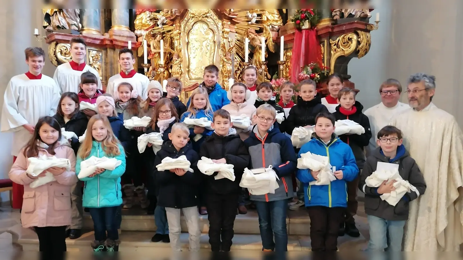 Die Erstkommunionkinder, die in den Händen ihre Albe halten, mit Ministranten, Pfarrer Helmut Brügel und Pastoralreferent Stefan Dotzler. (Bild: Anita Hartmann)