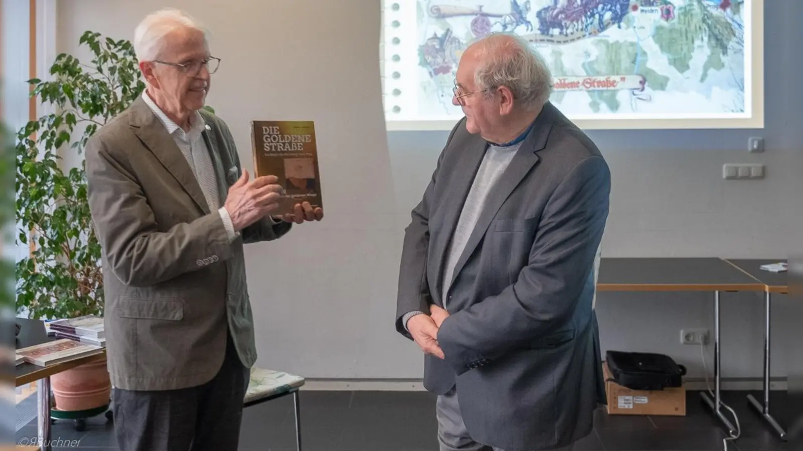 Rainer Christoph präsentiert dem Publikum und Pfr. Gerhard Pausch vom Verein Café Mitte e.V. sein neuestes Buch (Bild: Reinhold Buchner)