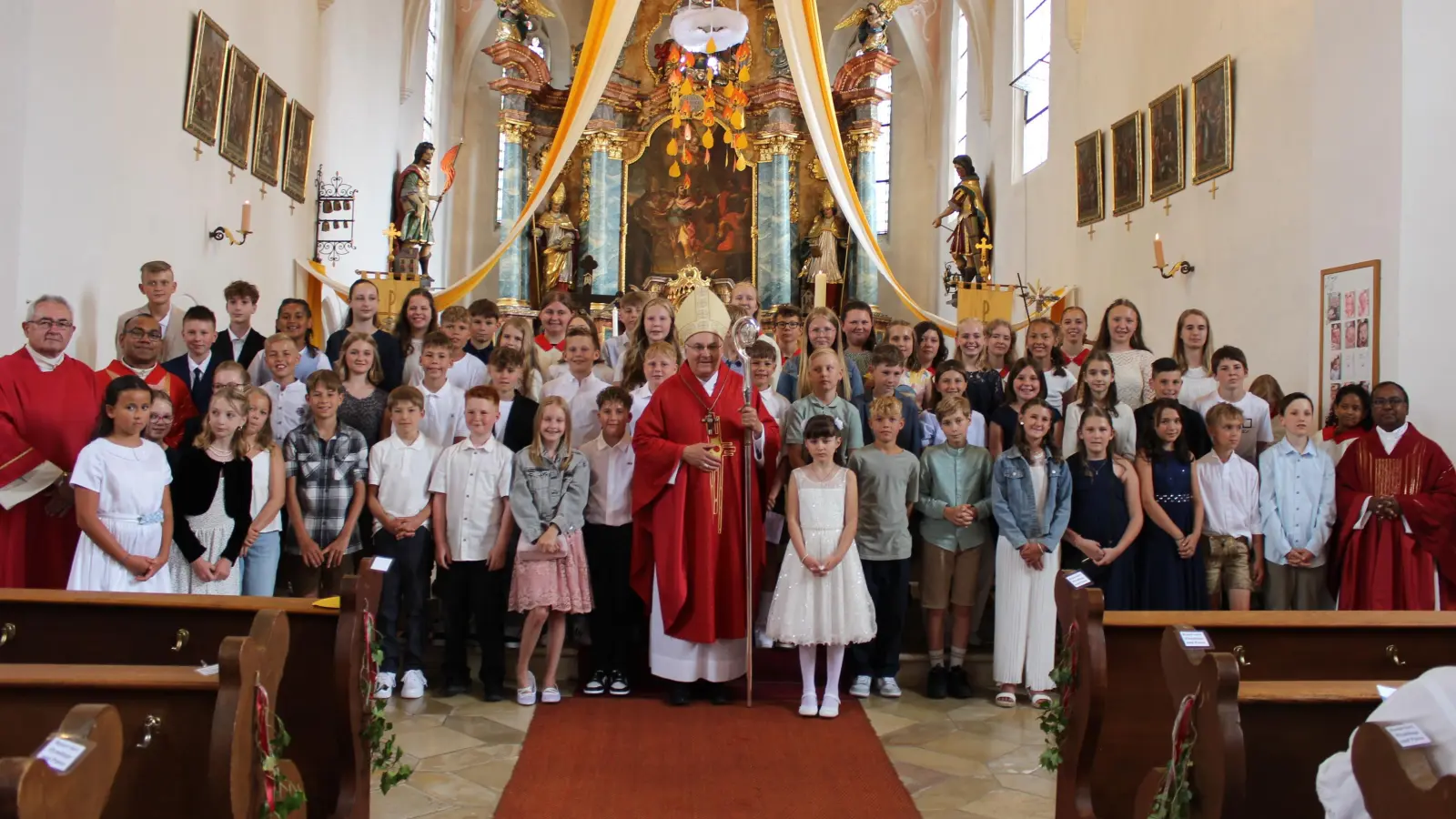 Nach dem Festgottesdienst stellte sich Bischof Dr. Rudolf Voderholzer inmitten der 52 Firmlinge zu einem Erinnerungsfoto in der Pfarrkirche St. Georg – unter den in der Firmvorbereitung angefertigten Feuerzungen mit den Namen der Jugendlichen. Links Diakon Dieter Gerstacker und Pfarrvikar Jimmy Joseph, rechts Stadtpfarrer Hruday Madanu. (Bild: Christian Trummer)