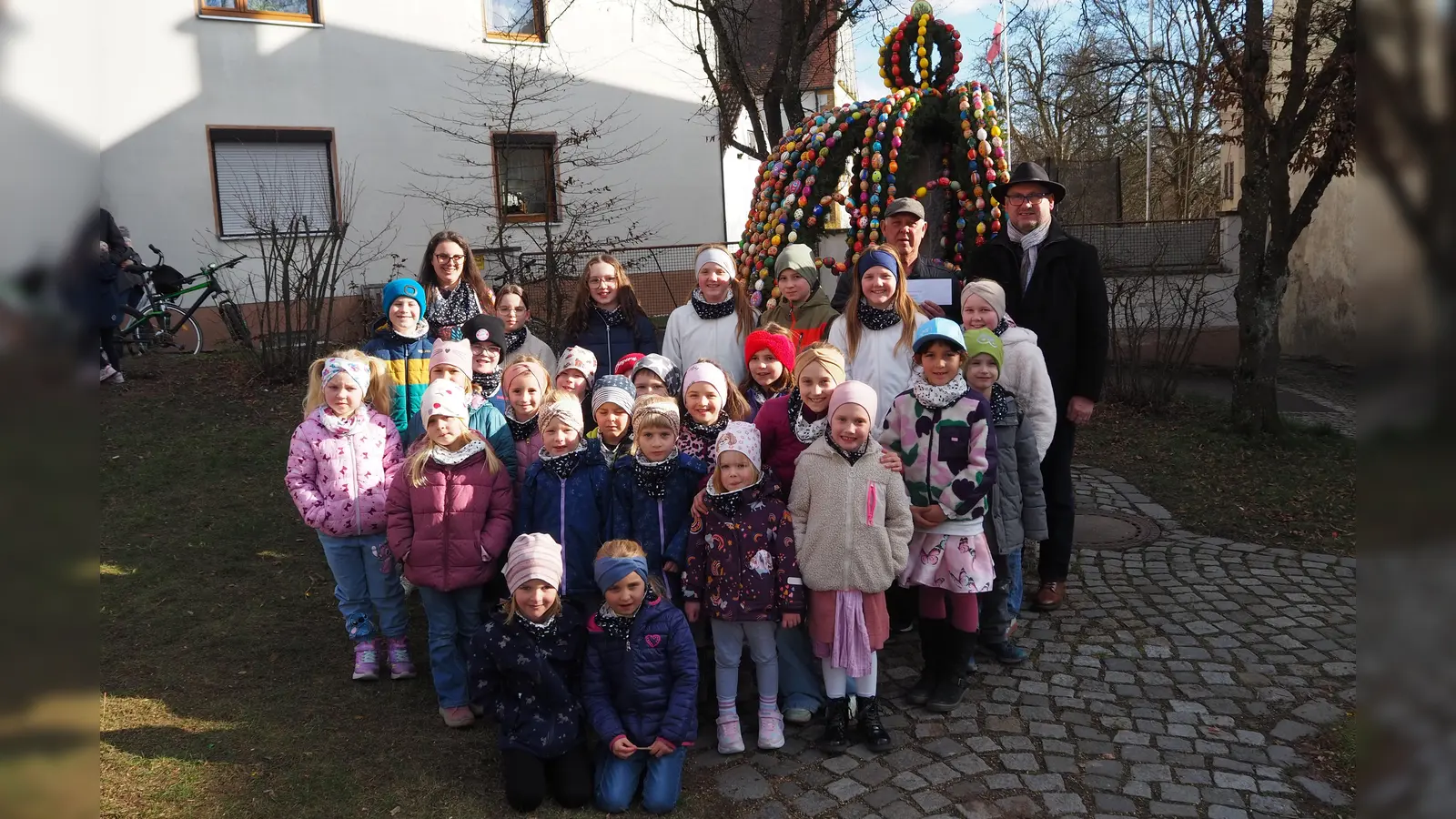 Mit Liedbeiträgen vor dem Osterbrunnen erfreute die Pfiffikusbande alle Anwesenden. Bürgermeister Richard Kammerer (hinten rechts) überreichte an Georg Seiser (hinten links) eine Geldspende. (Bild: Johann Kneißl )